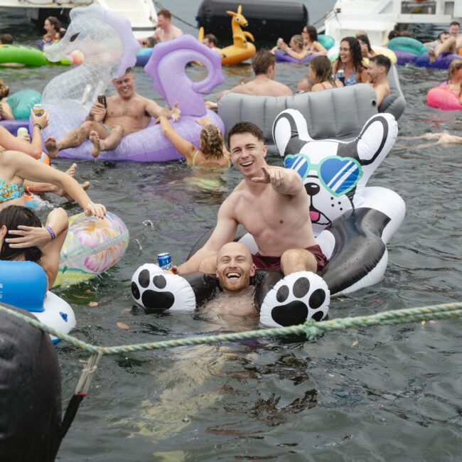A group of people enjoying a party on a river, floating on inflatable animals. Two men are in the foreground on a large inflatable cat, with others mingling nearby. Boats and a bridge are visible in the background.