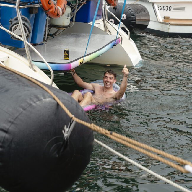Man lounging on an inflatable float in the water by a boat, giving a thumbs-up and smiling. There's a beverage can on the boat nearby. The scene is lively and joyful.