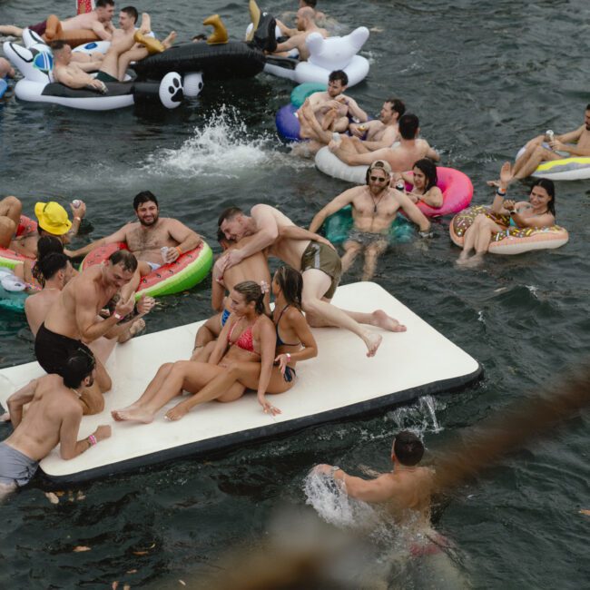 A lively scene of people enjoying a summer party on a lake. Various inflatable floats, including animals and a mattress, are occupied by smiling partygoers wearing swimsuits. The atmosphere is fun and carefree, with splashes and laughter in the water.