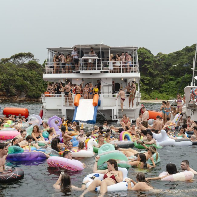A lively scene with people enjoying a party on inflatable floats in the water, near a large boat. The backdrop features lush greenery, and the boat has a slide into the water. The atmosphere is festive and crowded.