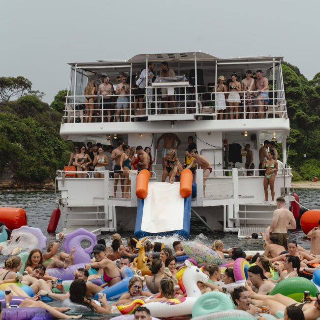 A large group of people enjoying a party on and around a two-story boat. Many are floating in the water on inflatable toys and others are on the boat deck. Trees are in the background, under an overcast sky.