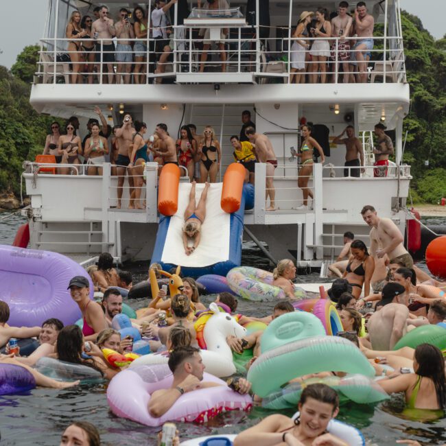 A crowded party scene on a boat with people celebrating and swimming. Many are using inflatable pool floats of various shapes and colors. One person slides into the water from a slide on the boat's deck.