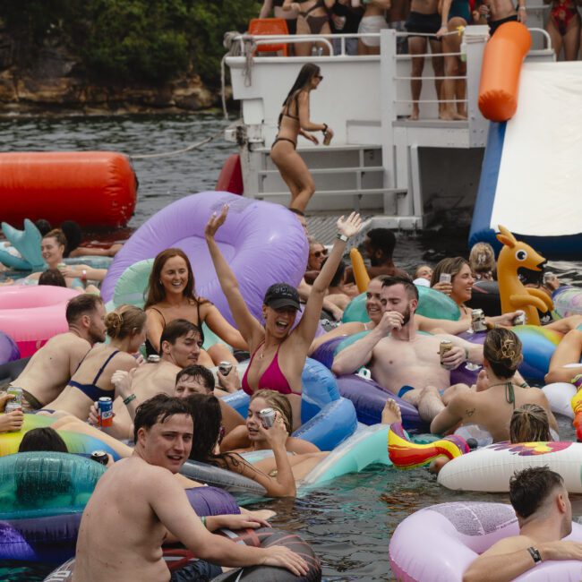 A group of people enjoy a party on water, floating on inflatable tubes near a boat. Many wear swimsuits and hold drinks, while some play on a slide attached to the boat. The atmosphere is lively and festive.