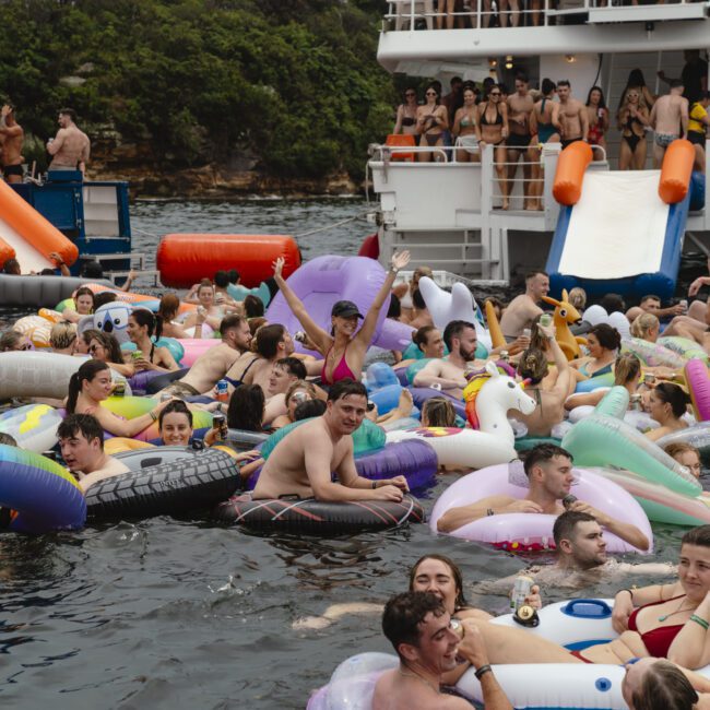 A large group of people enjoying a party on the water, floating with colorful inflatables near a boat. Some are on a slide leading into the water. The backdrop is a lush, green shoreline. The atmosphere is festive and lively.