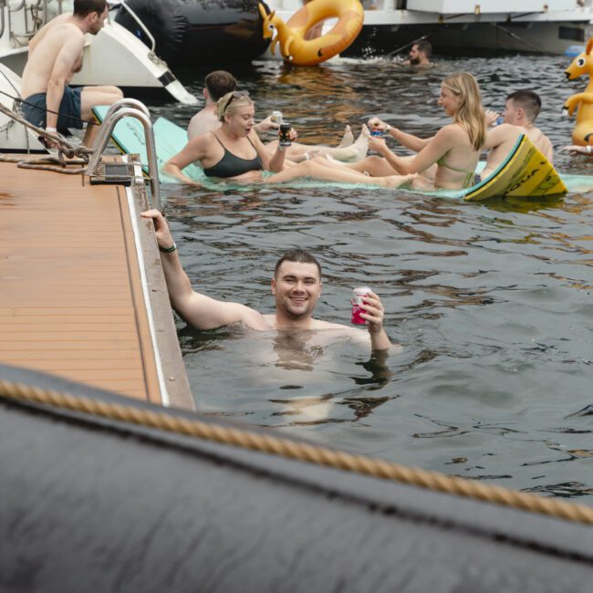 A group of people enjoys a pool party on a lake. Some are in the water holding drinks, while others relax on inflatables like pizza slices and ducks. There are boats in the background. The atmosphere is lively and fun.