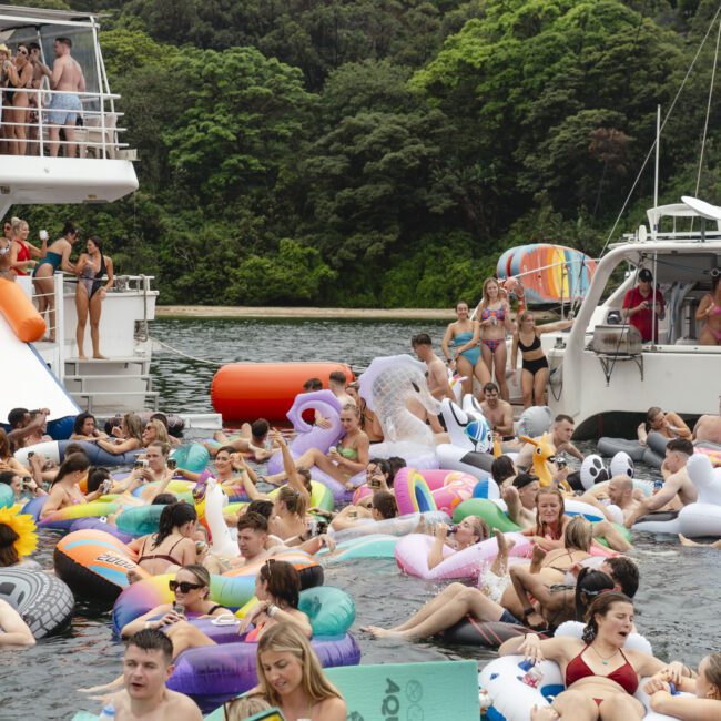 A large group of people enjoying a vibrant pool party on a lake. They are floating on colorful inflatable devices, with two boats nearby and lush green trees in the background.