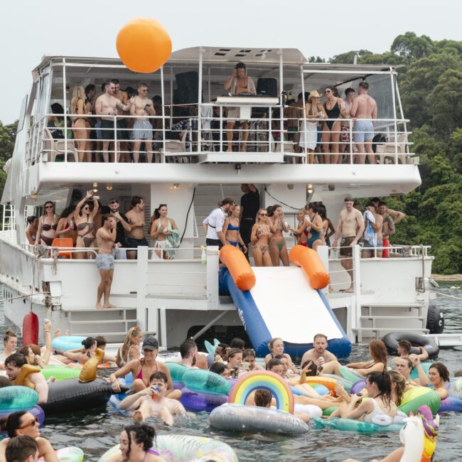 A large group of people enjoying a party on a double-deck boat. Many are in swimsuits, some are on the boat, while others are in the water on colorful inflatable floats. The backdrop features lush green trees.
