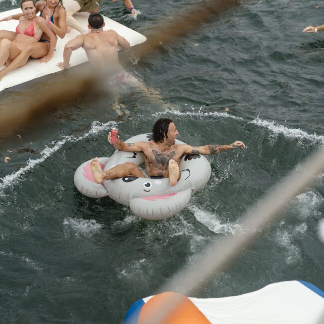 A group of people relax and play on large inflatable rafts and floaties in a body of water. They appear to be enjoying a sunny day, surrounded by rippling waves. One person is prominently lounging on a cute cloud-shaped float.