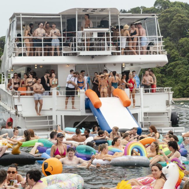 A lively boat party with people on a double-decker boat. Many are swimming in the water, using colorful inflatable floaties shaped like animals and objects. The scene is festive, with a natural backdrop of trees and greenery.