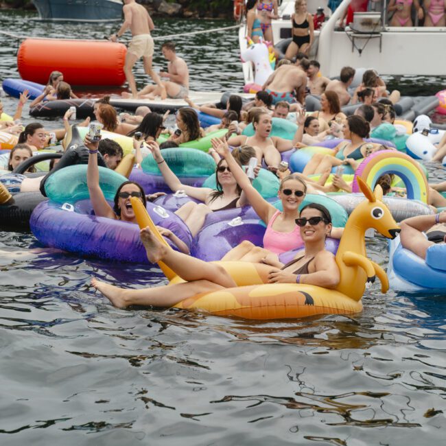 A lively scene of people enjoying a floating party on a body of water, with colorful inflatable rings and floats. Smiling individuals relax and take photos, while a boat in the background hosts more partygoers.