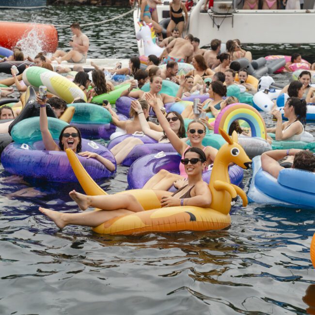 A lively group of people floating on colorful inflatable pool toys in the water. They're enjoying a sunny day, surrounded by a mix of animals and shapes, with a boat and more people in the background. A large orange buoy floats nearby.