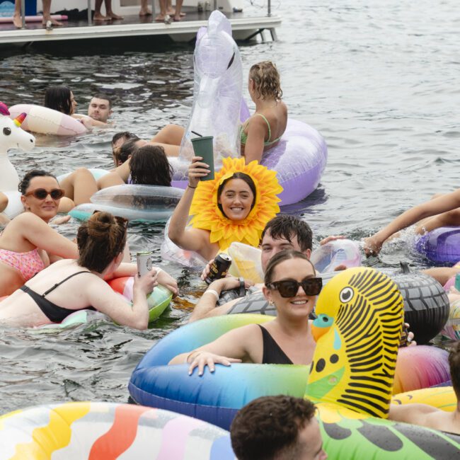 A large group of people enjoying a day on the water, floating on various inflatable toys. One person is wearing a sunflower costume and holding a drink, surrounded by others on unicorn and animal-shaped floats, with a boat in the background.