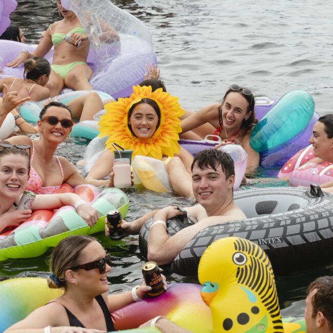 A group of people enjoying a lively day on a lake, surrounded by colorful inflatable rafts, including a parrot and a unicorn. One person wears a sunflower costume. Everyone appears relaxed and cheerful, holding drinks and laughing together.