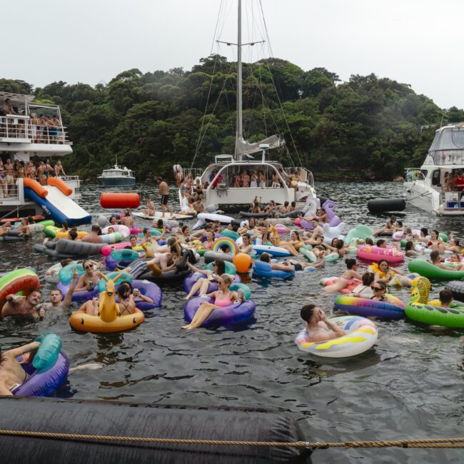 A lively scene of people enjoying a boat party on the water, surrounded by various colorful inflatable floats shaped like animals, fruits, and other fun designs. Several boats are docked nearby, and lush greenery can be seen in the background.
