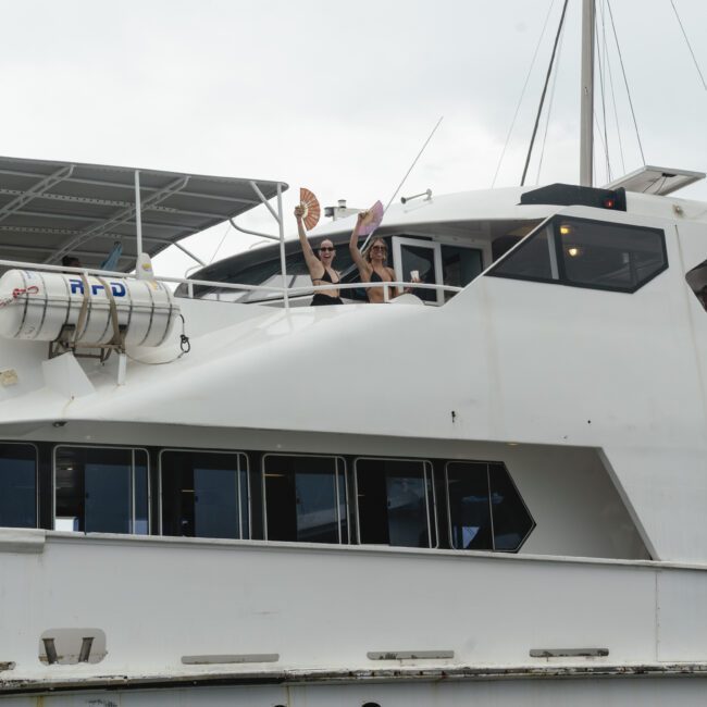 Two people waving from the upper deck of a large white yacht. The yacht has several windows and a visible lifebuoy. The sky is overcast.