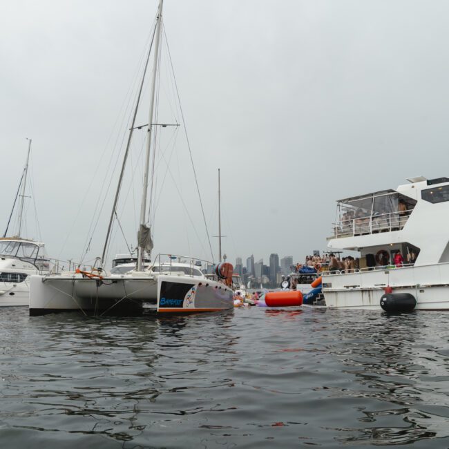 Boats gathered closely on a calm body of water with a city skyline in the hazy background. A lively crowd is visible on one of the larger boats, indicating a social event or party atmosphere. Overcast skies set a muted tone.