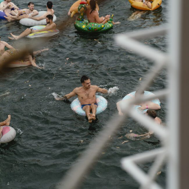People enjoying a swim in a river, lounging on colorful inflatable pool floats. The scene is viewed through the railing of a boat, adding a sense of perspective and movement. Some floats are shaped like animals, adding a playful touch.