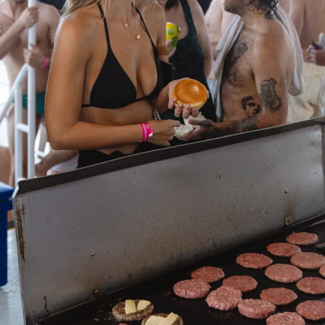 People are gathered around a grill on a boat. A woman in a black bikini holds a hamburger bun, while multiple patties and some steaks are cooking on the grill. The crowd appears relaxed and festive, wearing swimwear and enjoying the event.