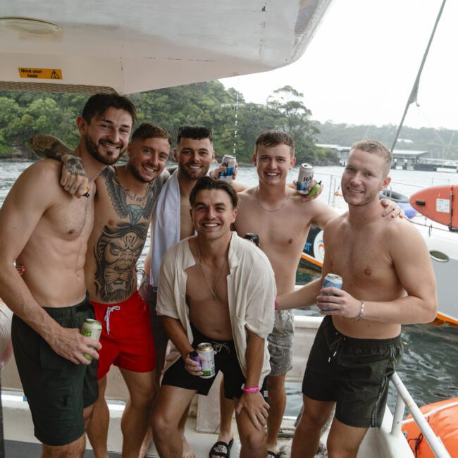 A group of six smiling people, some shirtless, standing on a boat. They are holding drinks and posing together. The background features water and trees under a cloudy sky.