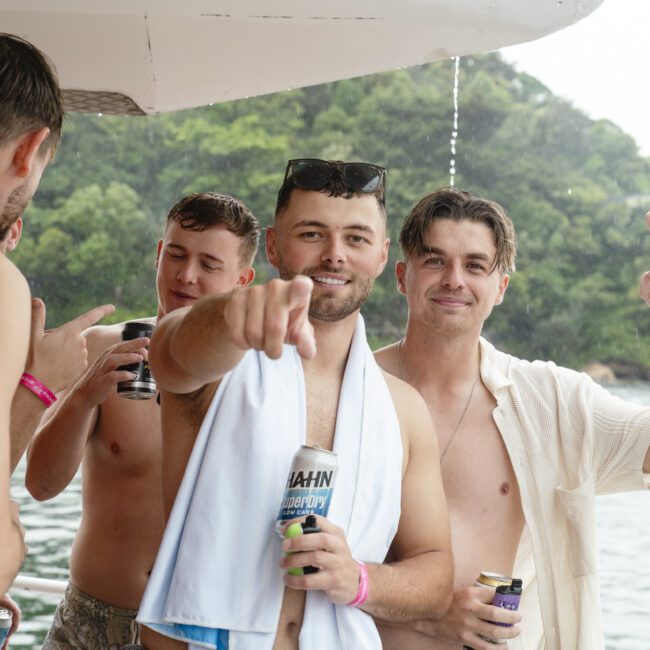 A group of shirtless men on a boat, some with drinks in hand. One man points at the camera while another flashes a peace sign. They are wearing pink wristbands. The background shows a body of water and trees.