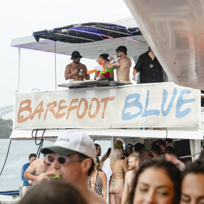 A lively boat party with people dancing and socializing. The boat has a sign that reads "Barefoot Blue." Two DJs are on an elevated deck playing music, while attendees enjoy the atmosphere. In the background, a glimpse of a bridge is visible.
