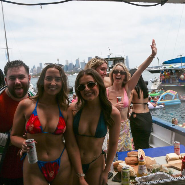 A group of people in swimsuits enjoy a boat party. Two women in bikinis pose in the foreground, smiling. Other individuals are behind them, with some waving. The city skyline and boats are visible in the background. Food and drinks are on the table.