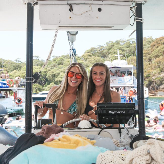 Two women in swimwear are smiling at the helm of a boat, surrounded by colorful towels and clothes. Other boats and people in swim rings are visible in the background on a sunny day.
