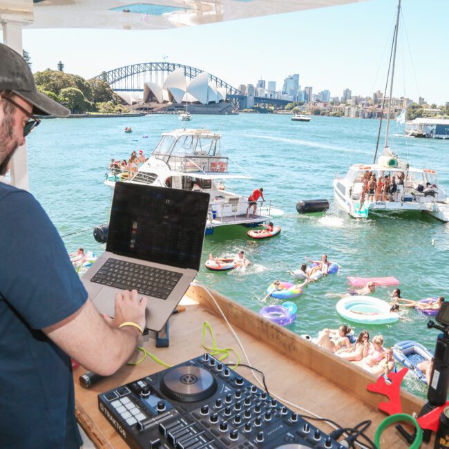 A DJ plays music on a boat overlooking a vibrant outdoor party. People are swimming and relaxing on inflatables in the water. In the background, the Sydney Harbour Bridge and Opera House are visible under a clear blue sky.