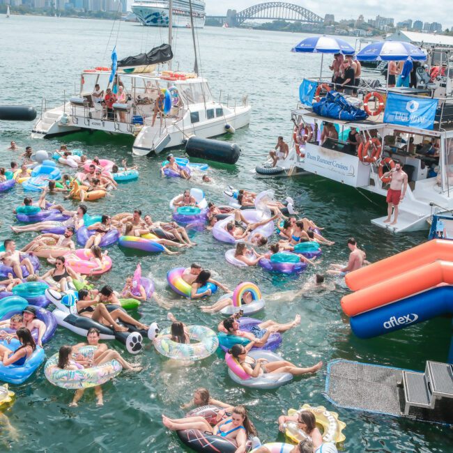 A lively scene on water shows numerous people enjoying themselves on colorful inflatable tubes. They are surrounded by boats with more people watching and participating. The backdrop features a bridge under a partly cloudy sky.