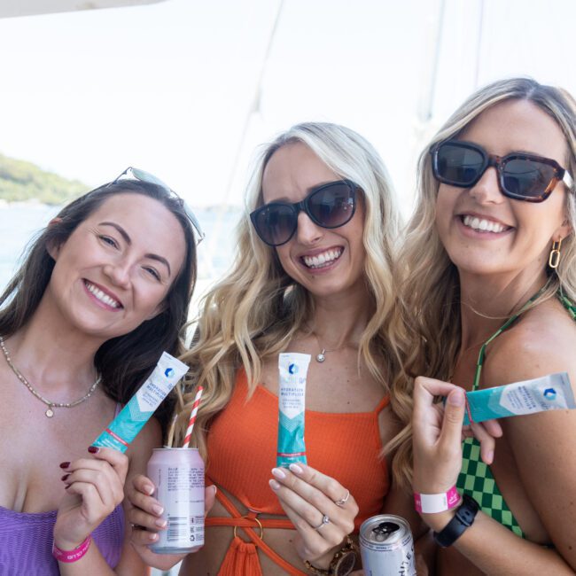 Three women in sunglasses smile while holding beverage cans and tubes on a boat. They stand close together against a backdrop of water and trees, enjoying a sunny day.