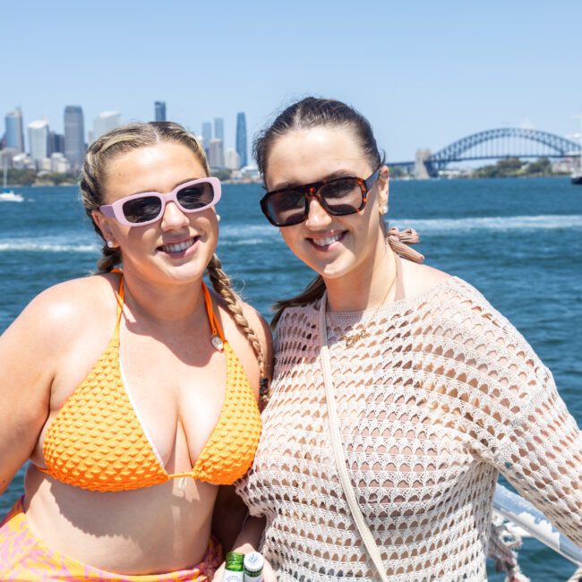 Two women posing and smiling on a boat, with a city skyline and a large bridge visible in the background. One wears an orange bikini top, the other a white crocheted top. The sky is clear and sunny.