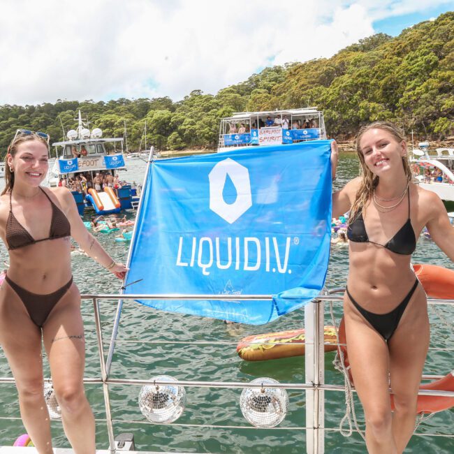 Two smiling women in bikinis stand on a boat holding a blue Liquid I.V. banner. Behind them, other boats are anchored, and people are swimming and enjoying the sunny day on the water surrounded by green trees.