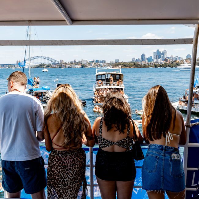 Four people on a boat enjoying a sunny day, facing a view of multiple boats and people swimming in a harbor. The city skyline and iconic bridge are visible in the background.