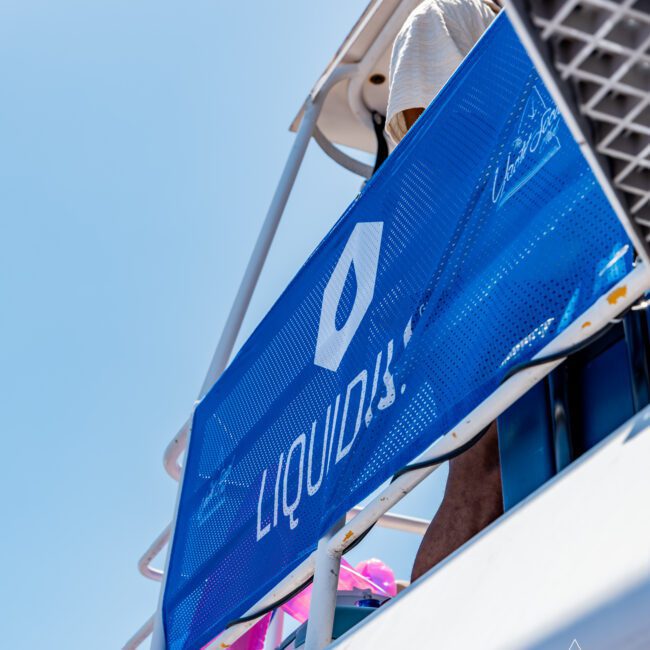 Close-up of a blue banner with the word "LIQUID" on a boat railing. The sky is clear and blue, and part of the boat structure is visible. The logo and text "The Yacht Social Club" appear in the bottom corner.