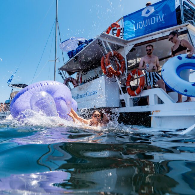 A person swimming near a boat, holding a large purple inflatable float. Other people are on the boat, which has multiple life rings and umbrellas on deck. The boat displays "Rum Runner Cruises" and "Liquid Liv" branding.
