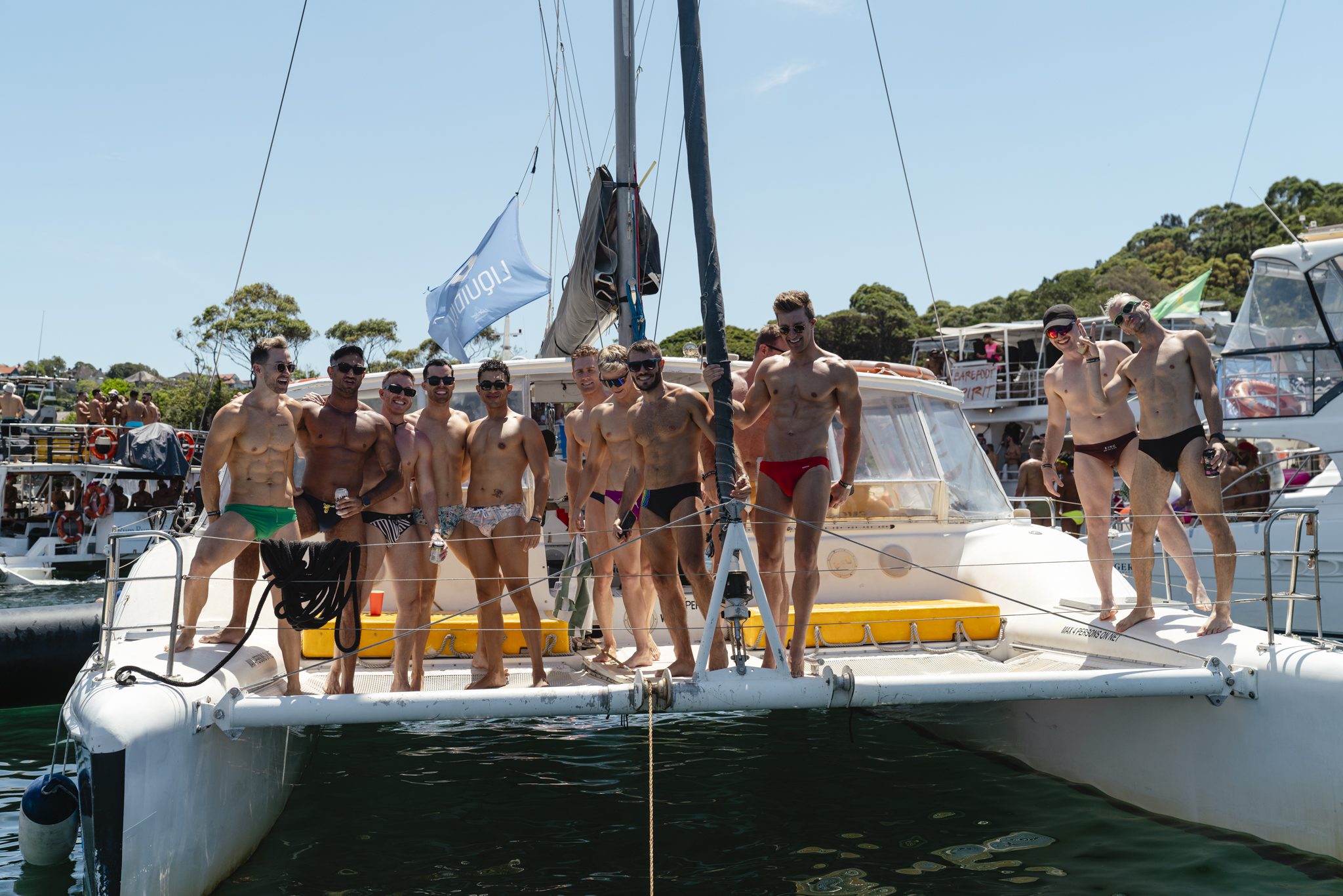 A group of people wearing swimsuits stand on the front of a catamaran in a sunny setting. There are other boats and people visible in the background, with trees and a clear blue sky overhead.