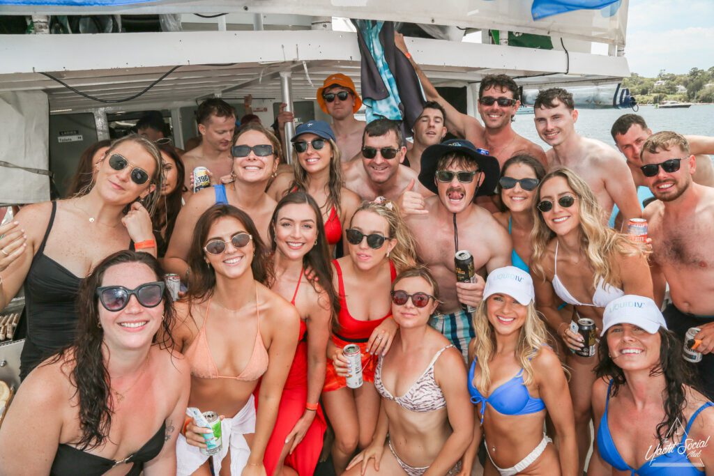 A large group of people dressed in swimwear is gathered on a boat, smiling and holding drinks. They appear to be enjoying a sunny day on the water, with some wearing hats and sunglasses.