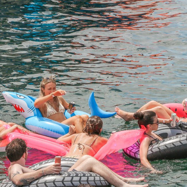A group of people relaxing on colorful inflatables in the water. Some hold drinks while lounging on pink and gray tubes, and one inflatable resembles a shark. The scene conveys a fun, leisurely gathering.
