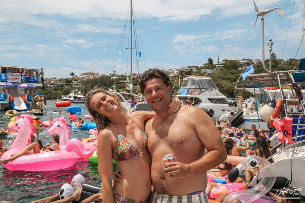 A smiling man and woman in swimwear stand on a boat, with numerous inflatable floaties and people in the water around them. The scene includes several anchored boats, a clear blue sky, and coastal greenery in the background.
