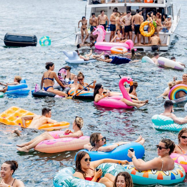 People are enjoying a sunny day on the water, lounging on colorful inflatables of various shapes like flamingos and unicorns. A boat is nearby with more people on board, and the background shows a distant shoreline and buildings.