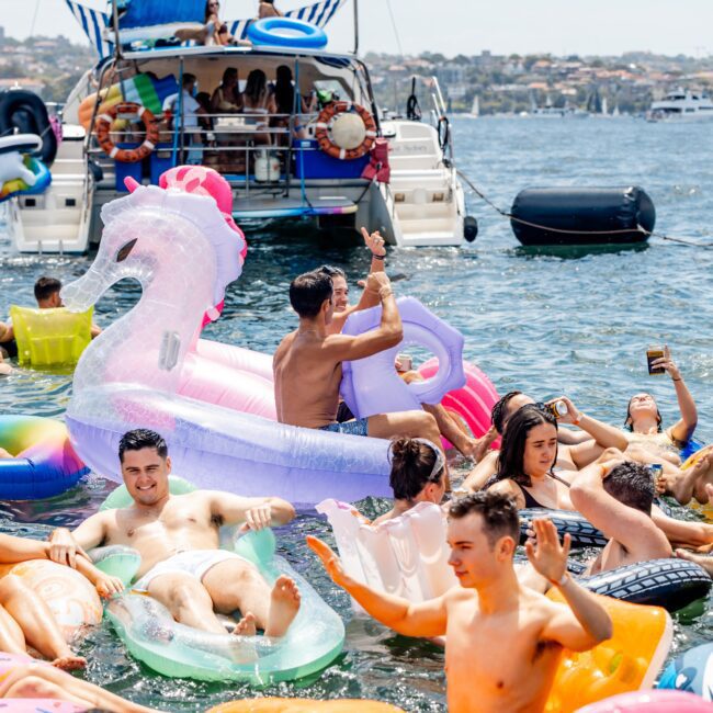 A group of people enjoying a sunny day, floating on colorful inflatables in the water near moored boats. Visible are a unicorn float, several pool rings, and people smiling and waving, with a cityscape in the background.