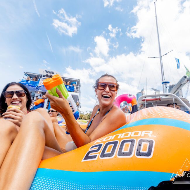 A group of people laughing and relaxing on a colorful inflatable raft in the water. The sky is clear and blue, with surrounding boats in the background. One woman holds a drink container, and everyone appears to be enjoying a sunny day.