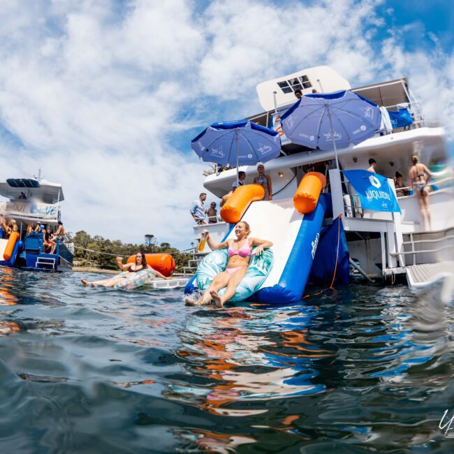 A group of people enjoying a sunny day on a yacht, with some relaxing on inflatable floats and others using a water slide attached to the boat. Bright blue sky and city buildings visible in the background.