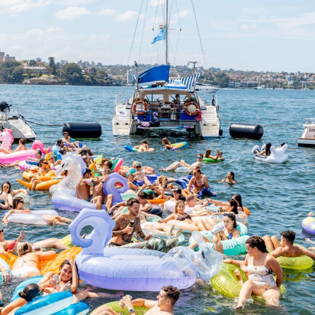 A group of people enjoying a sunny day on colorful inflatable tubes in the water. A boat is anchored nearby, and the shoreline with greenery is visible in the background. The atmosphere is lively and festive.