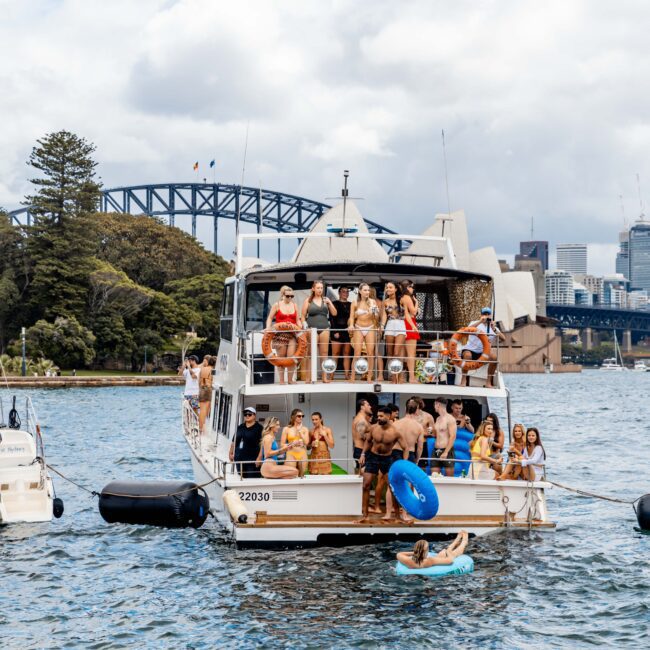 A group of people enjoying a party on a yacht in a harbor, with city buildings and a large bridge visible in the background. Inflatable toys float nearby on the water, and the sky is partly cloudy.