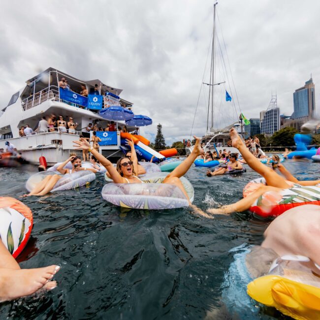 People enjoying a party in the water, lounging on inflatables with arms raised. A large yacht in the background has people on board. Skyscrapers and a cloudy sky are visible in the distance.