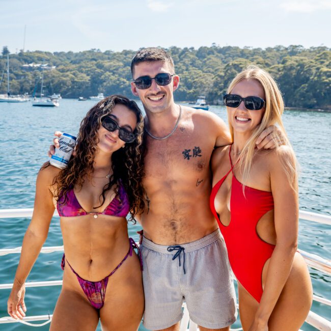 Three people in swimwear stand on a yacht with water and forested land in the background. The two women wear sunglasses and swimsuits, while the man in the middle is shirtless, holding a drink. The setting appears sunny and festive.