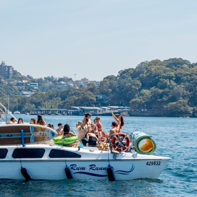 A white boat named "Rum Runner" on the water, carrying a group of people enjoying a sunny day. They are gathered on the deck with city buildings and lush green trees visible in the background. The water is calm and the sky is clear.
