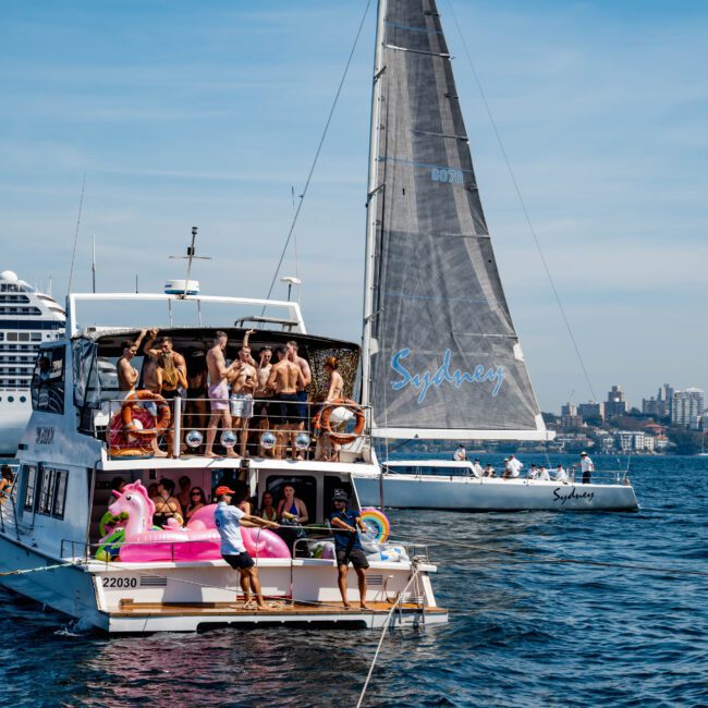 A group of people enjoying a lively gathering on a yacht adorned with colorful inflatables. In the background, a sailboat named "Sydney" is sailing on a sunny day. The scene is set against a cityscape and a large cruise ship.