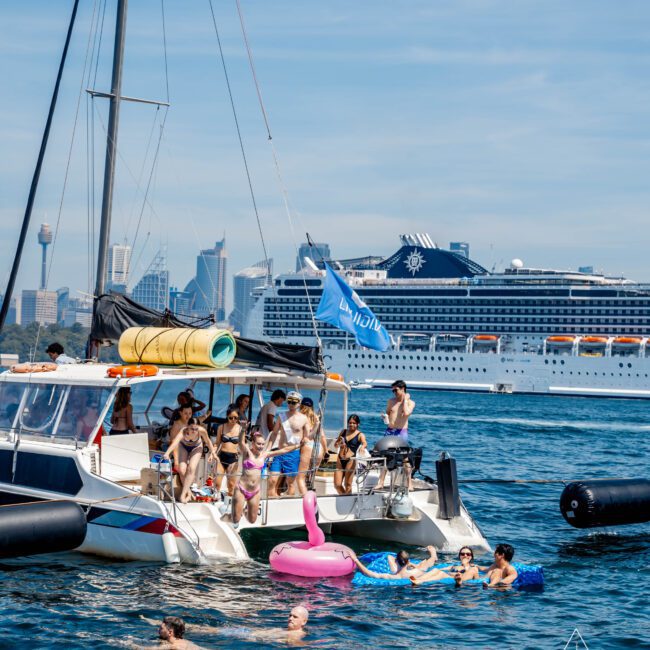 A group of people enjoying a sunny day on a sailboat and in the water, with a pink flamingo float nearby. A large cruise ship and city skyline are visible in the background under a clear blue sky.